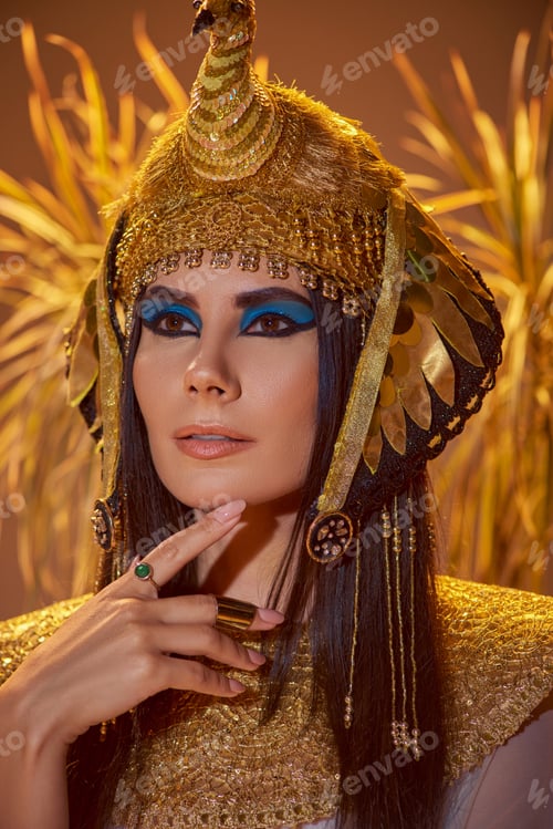 Preview: Elegant woman in egyptian look and headdress posing near blurred plants on brown background