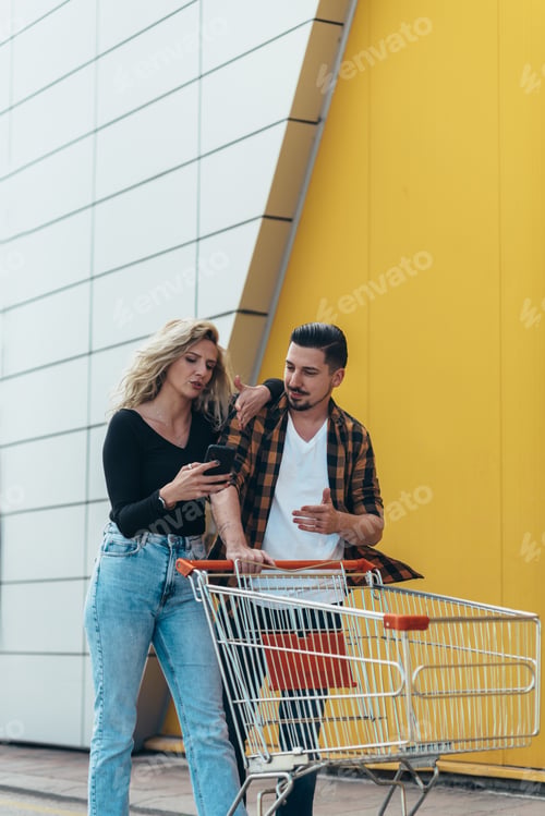 Preview: Young beautiful couple using smartphone while holding a supermarket shopping cart
