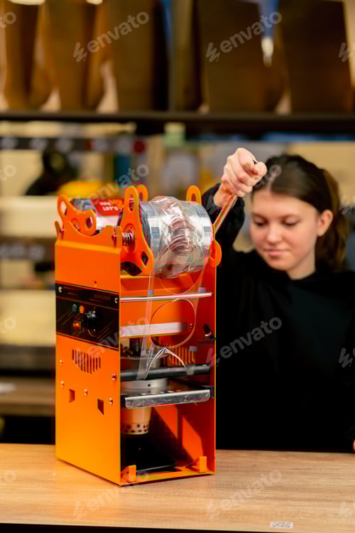 Preview: close-up of an orange machine packaging drinks with plastic on a bar counter girl using the machine