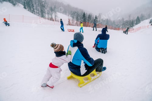 Preview: Small child pushes his dad on a sled down a snowy hill. Back view
