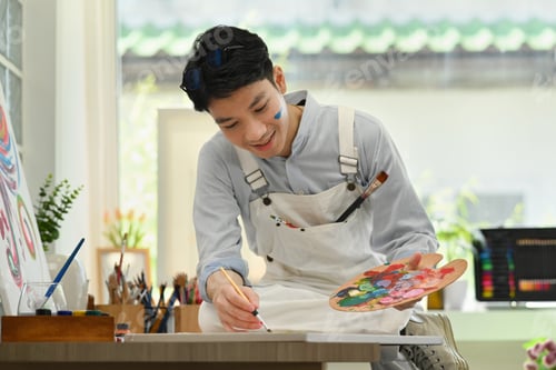 Preview: Happy asian male artist sitting in bright art workshop and enjoy creating his artwork.