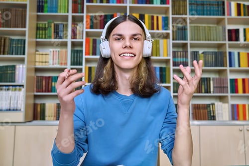 Preview: Teenage college student in headphones looking at webcam, sitting at desk in library