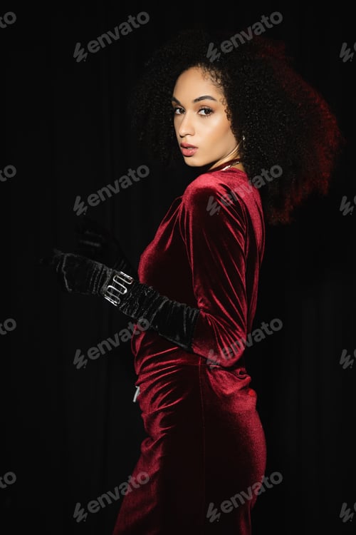 Preview: Trendy african american woman in velvet burgundy dress and glove looking at camera isolated on black