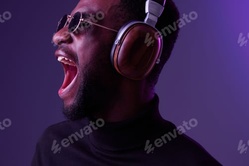Preview: Young African man shouting joyfully while wearing stylish headphones and sunglasses against a