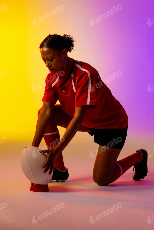 Preview: African american female rugby player crouching with rugby ball over neon pink lighting