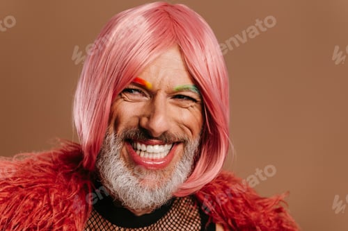 Preview: Happy mature gay man with rainbow colored eyebrows wearing pink wig against brown background