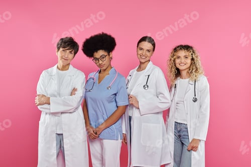 Preview: cheerful and multiethnic doctors smiling at camera isolated on pink, breast cancer concept
