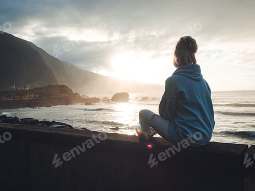 Preview: Blonde girl wearing a hoodie, sitting near the ocean enjoying the view during sunset