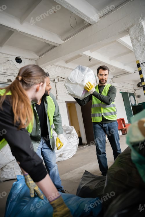 Preview: Worker in protective vest and glove holding plastic bag with trash near blurred interracial