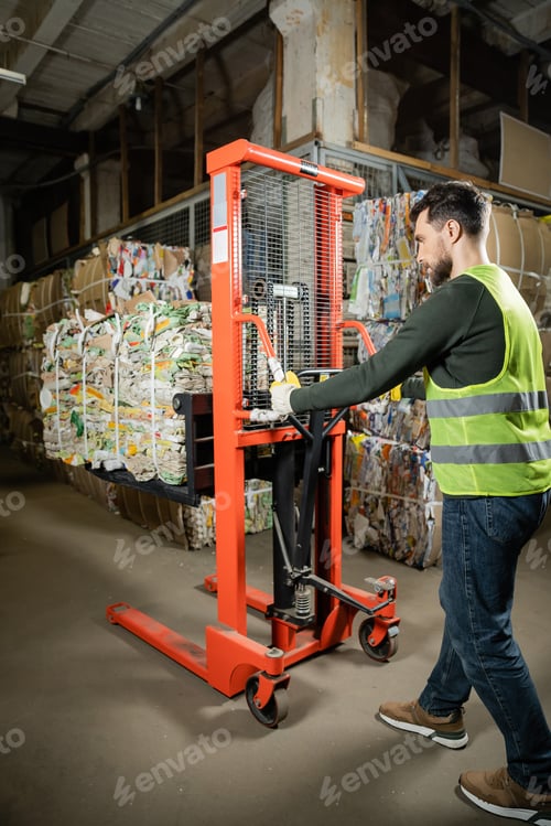 Preview: Side view of bearded sorter in protective vest and gloves using hand pallet truck with waste paper