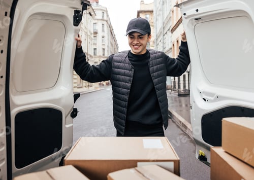 Preview: Smiling man in uniform and cap looking at cardboard boxes while standing at van trunk