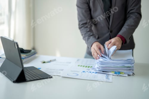 Preview: A man is standing in front of a table with a pile of papers and a laptop