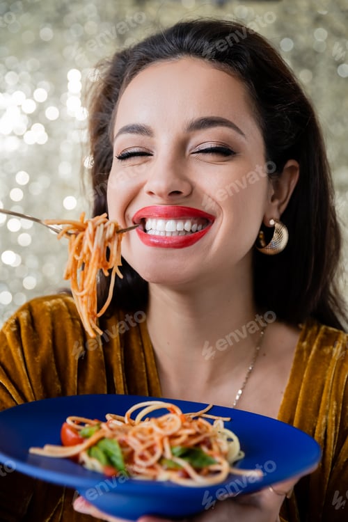 Preview: cheerful brunette woman holding blue plate and fork with pasta on shiny background