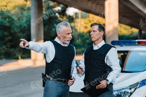 Preview: mature male police officer pointing by finger to colleague with clipboard near car at street