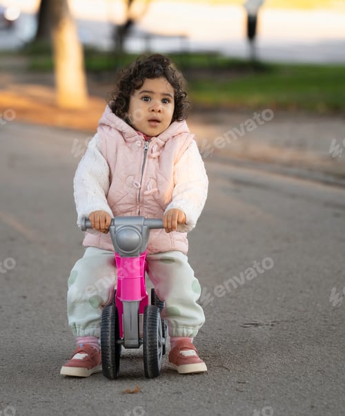 Preview: Cute toddler hispanic girl riding a pink balance bike in the park