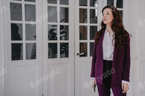 Preview: Attractive tired businesswoman holding phone looking aside dressed in burgundy suit at office
