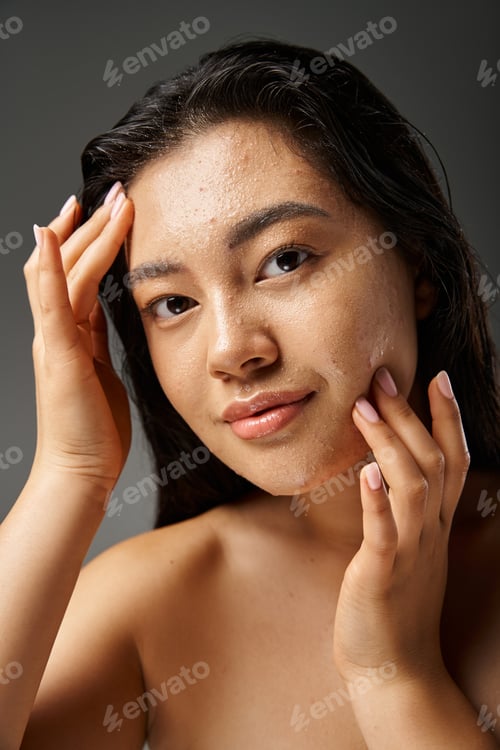 Preview: young asian woman with brunette hair toughing wet face with acne and looking at camera on grey