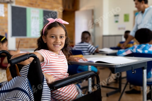 Preview: Portrait of happy biracial school girl sitting in wheelchair in class at elementary school