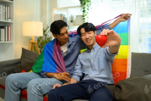 Preview: Affectionate gay couple sitting on a couch holding rainbow flag and red heart