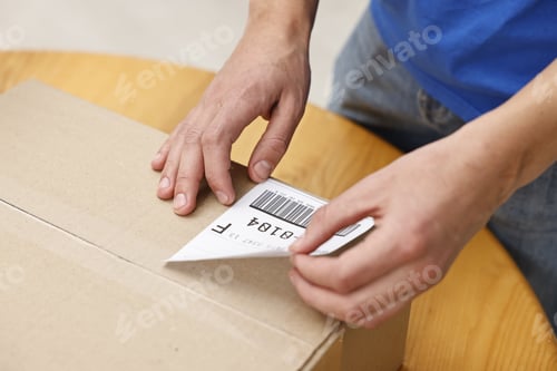 Preview: Man sticking shipping label with barcode on parcel at wooden table indoors, closeup