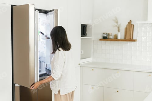 Preview: Stylish woman opening fridge and looking inside in new minimal white kitchen