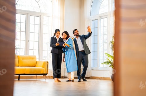 Preview: Indian woman real estate agent with young married couple walking in new property with tablet