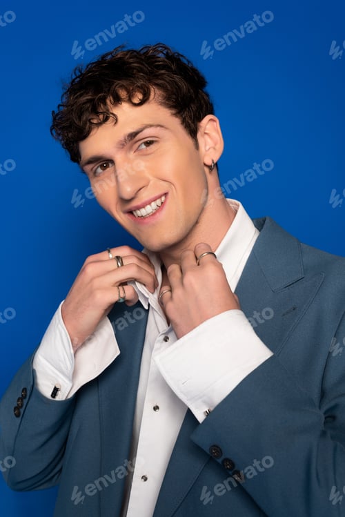 Preview: Portrait of cheerful curly man in jacket adjusting shirt isolated on blue