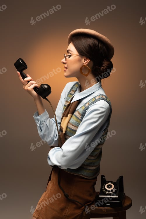 Preview: stylish young woman in beret and sunglasses looking at vintage telephone on brown