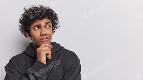 Preview: Horizontal shot of pensive curly haired teenage man keeps hand on chin concentrated aside thinking