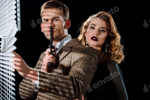 Preview: selective focus of handsome gangster holding gun near face and touching window blinds near woman