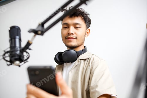 Preview: Smiling Broadcaster Using Smartphone in Front of Microphone at Home Studio