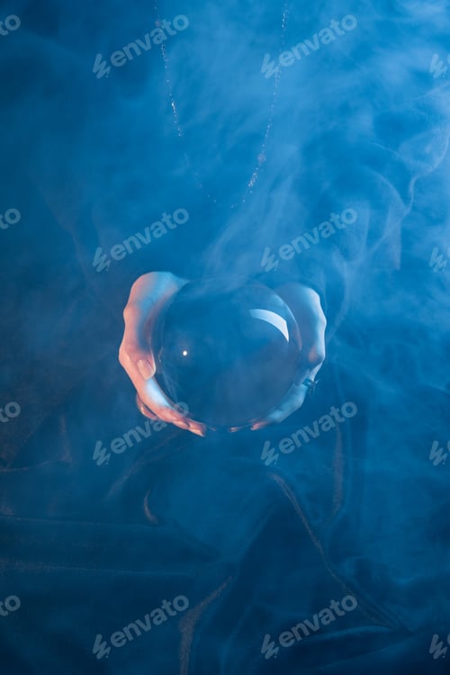 Preview: Cropped view of witch holding crystal ball above table on dark blue background
