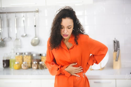 Preview: woman with curly hair standing in modern kitchen taking support of counter holding her abdomen hard