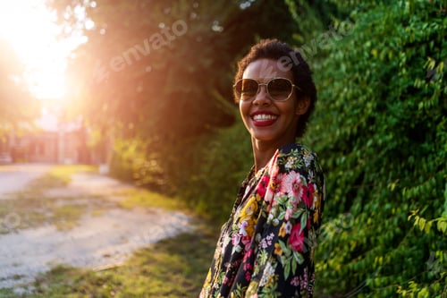 Preview: Portrait of happy young woman wearing sunglasses outdoors at sunset