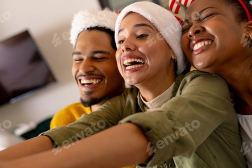Preview: Young multiracial friends wearing Santa hats laughing and taking selfie at Christmas party, at home