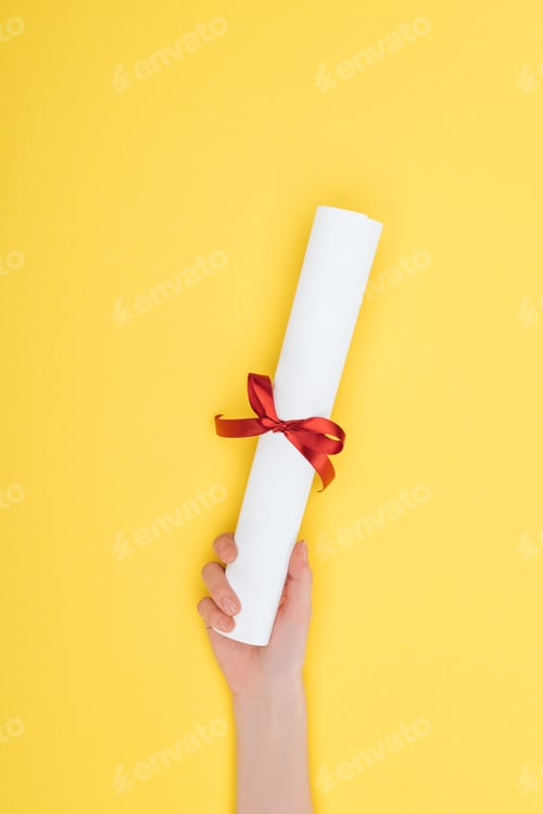 Preview: Cropped view of woman holding diploma with ribbon on yellow surface