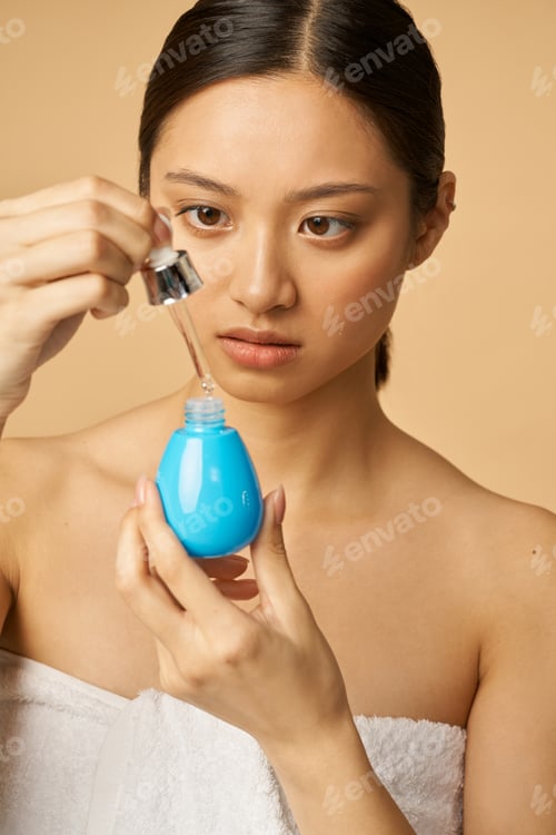 Preview: Portrait of young woman looking focused while holding oil drop serum, posing isolated over beige