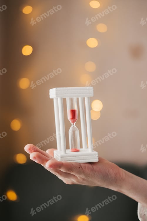 Preview: hands hold a white hourglass on the background of a Christmas garland. Hourglass close-up.