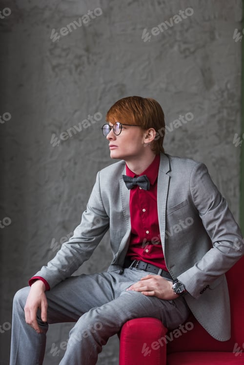 Preview: Profile of male model in eyeglasses dressed in suit sitting on edge of couch