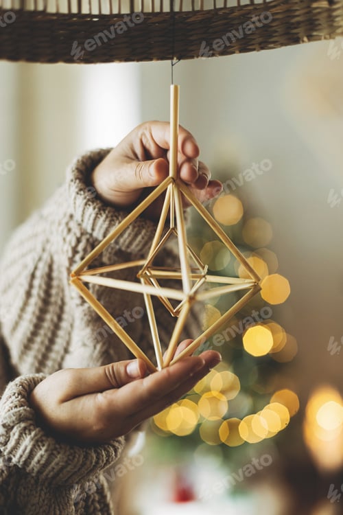 Preview: Hands in cozy sweater hanging rustic nordic straw decoration on background of festive golden lights