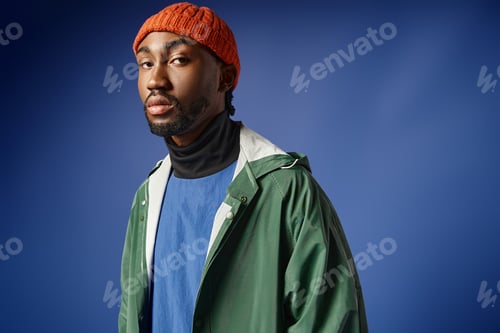 Preview: stylish african american man in knitted hat and trendy winter attire posing on blue backdrop