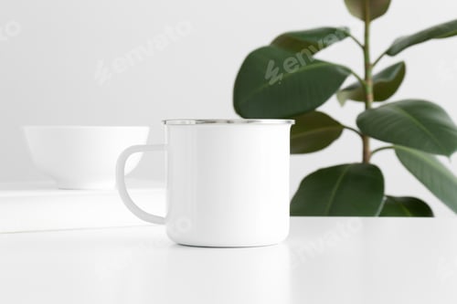 Preview: Enamel mug mockup with workspace accessories on a white table and a ficus plant.