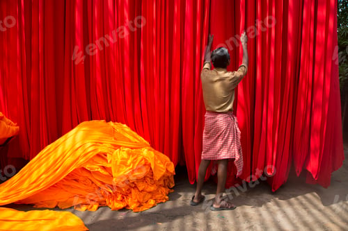 Preview: Rear view of man hanging up vibrant and bright red fabric, heap of orange fabric on floor.