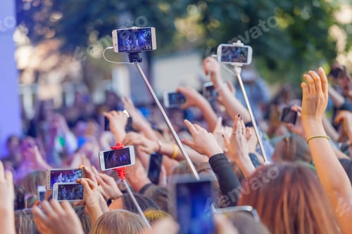 Preview: Teenagers using their phones at the music concert, hands up. Mobiles with selfie sticks.