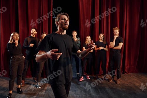 Preview: Man standing against people. Group of actors in dark colored clothes on rehearsal in the theater