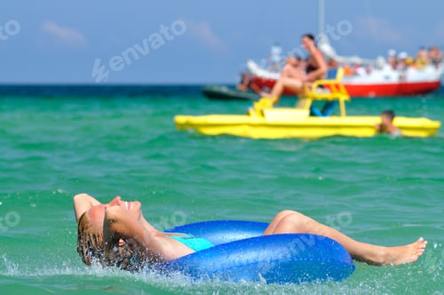 Preview: Young blond woman in blue bikini riding on swimming circle in sea and smiling