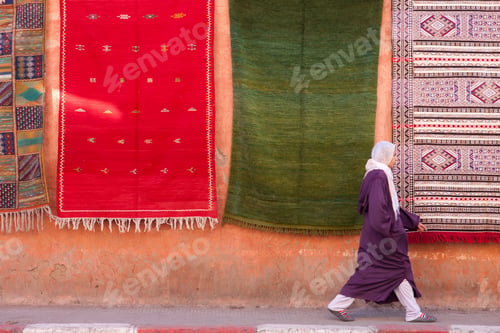 Preview: An elderly person walking past vibrant hanging rugs on an orange wall