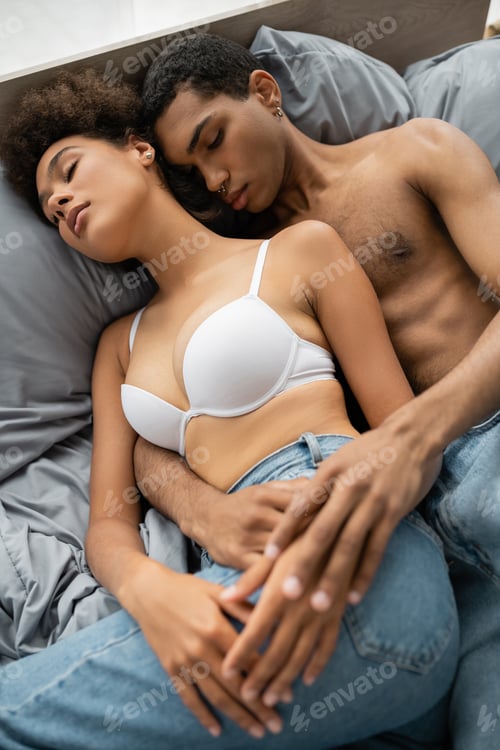 Preview: high angle view of shirtless african american man and woman in white bra lying on bed with closed