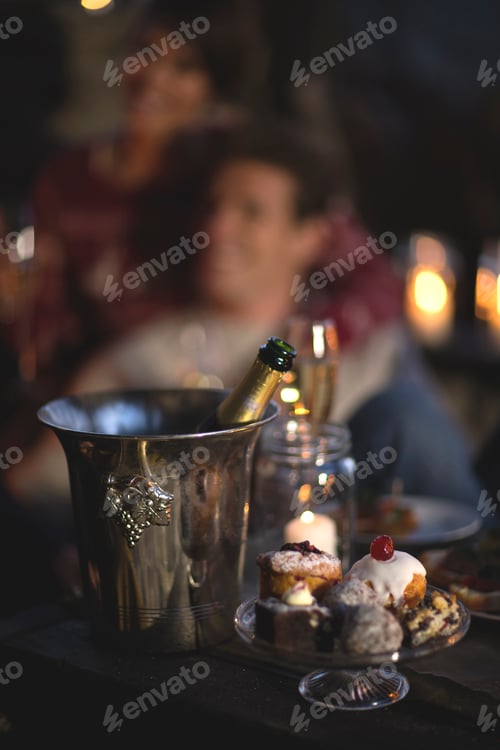 Preview: Close up of champagne bottle in cooler, cakes on a stand, person in the background.
