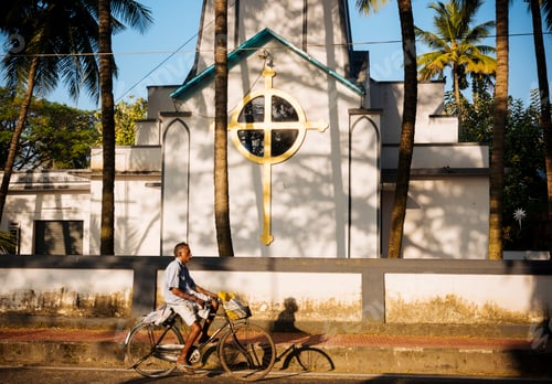 Preview: A man rides a bicycle past a white church with a golden cross in the warm light of the afternoon.